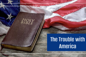 Holy Bible and an American flag on a rustic wooden table with text that reads, The Trouble with America